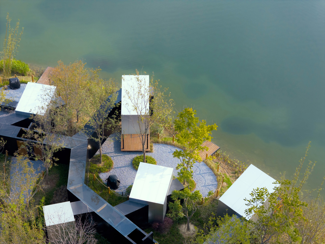 Top-down view of circular courtyard with gravel paths and planted trees between angled pavilions at water's edge