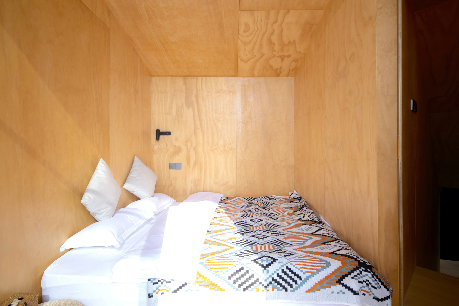 Plywood-clad sleeping room with white bedding and patterned throw under natural daylight