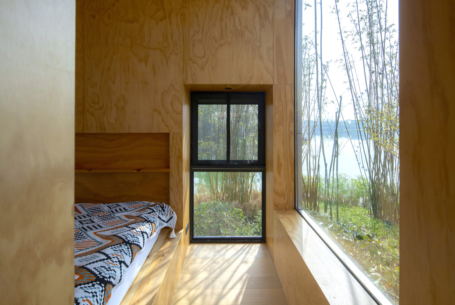 Plywood-lined sleeping alcove with floor-to-ceiling window overlooking bamboo groves and water