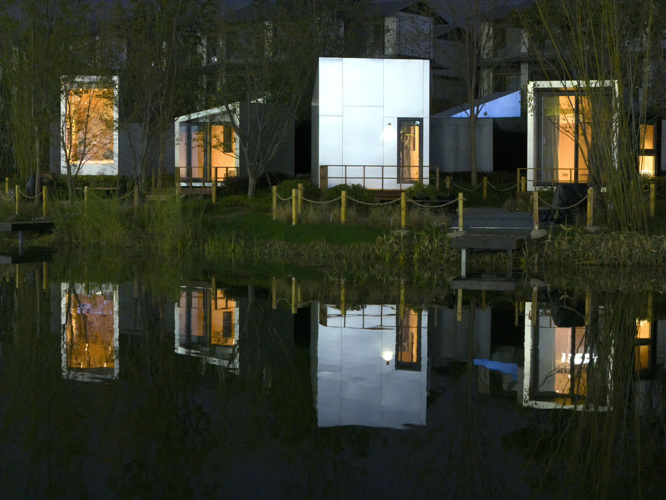 Illuminated pavilions with curved and cubic forms mirrored in a pond at twilight