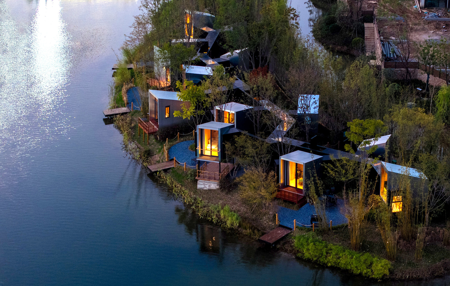Aerial view of clustered pavilions with illuminated glazing nestled among dense vegetation along a waterfront at dusk