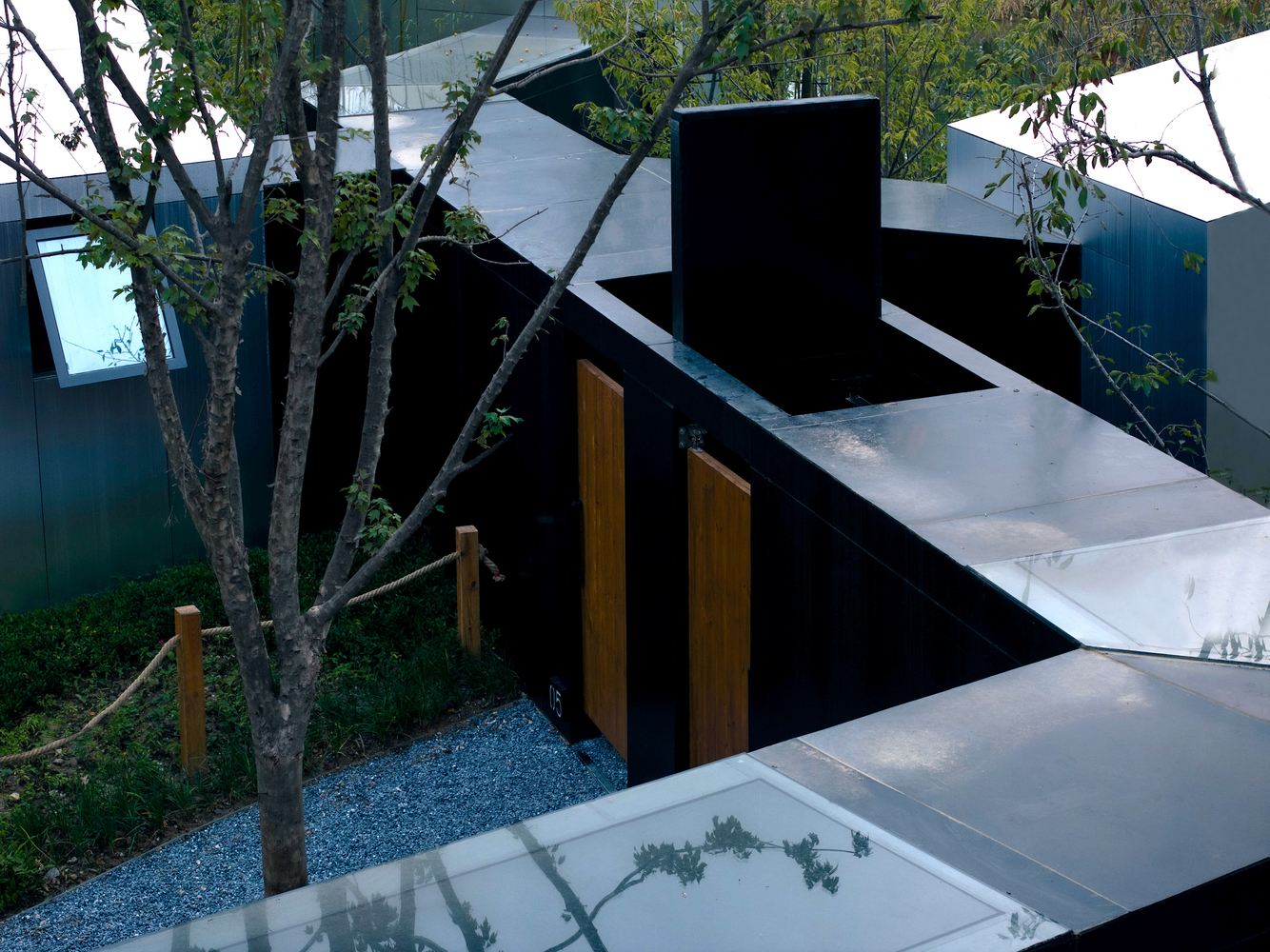 Overhead view of black steel volumes with timber doors arranged around a courtyard tree and gravel pathway