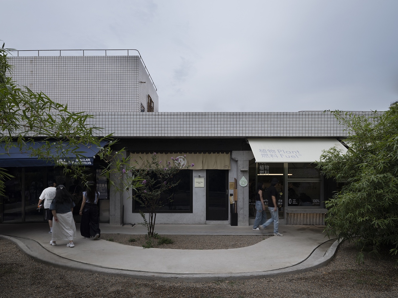 Street-level facade with white mosaic tile upper volume and retail storefronts beneath shallow canopy at dusk