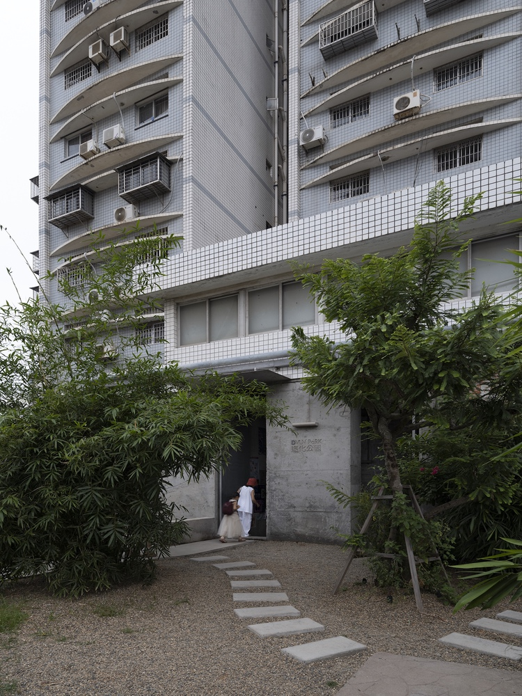 Stepping stone path through planted garden leading to concrete doorway beneath white tiled residential facade