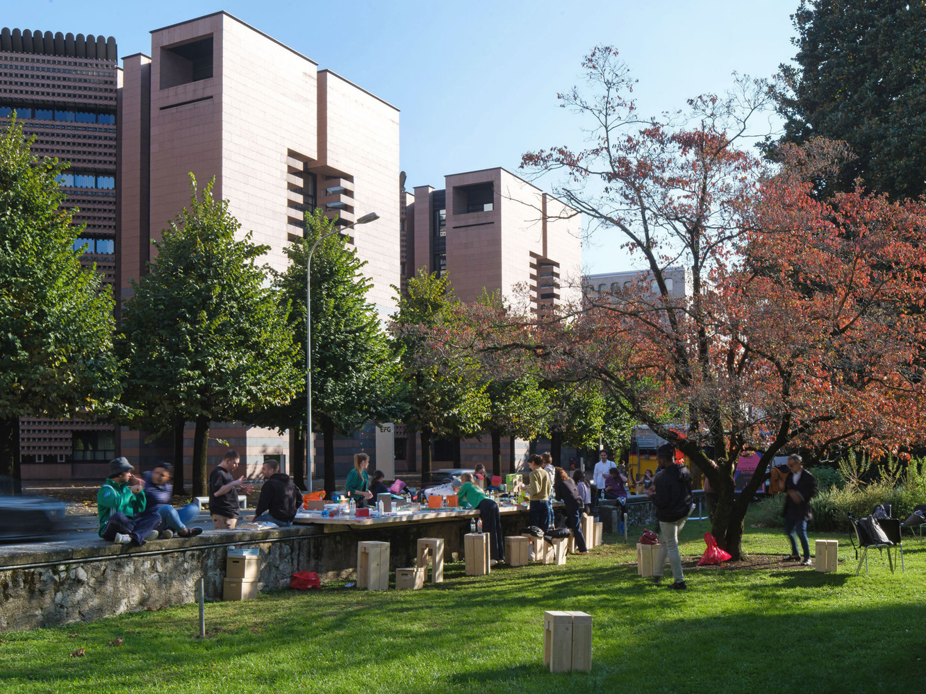 People gathered around a long timber table in a park with pink masonry buildings behind