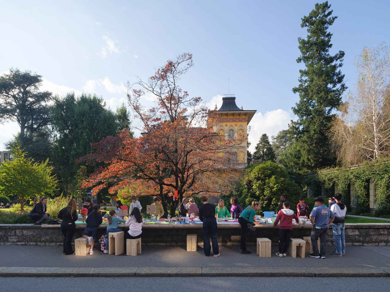 Long timber table with people seated on block stools beneath an autumn tree in a park