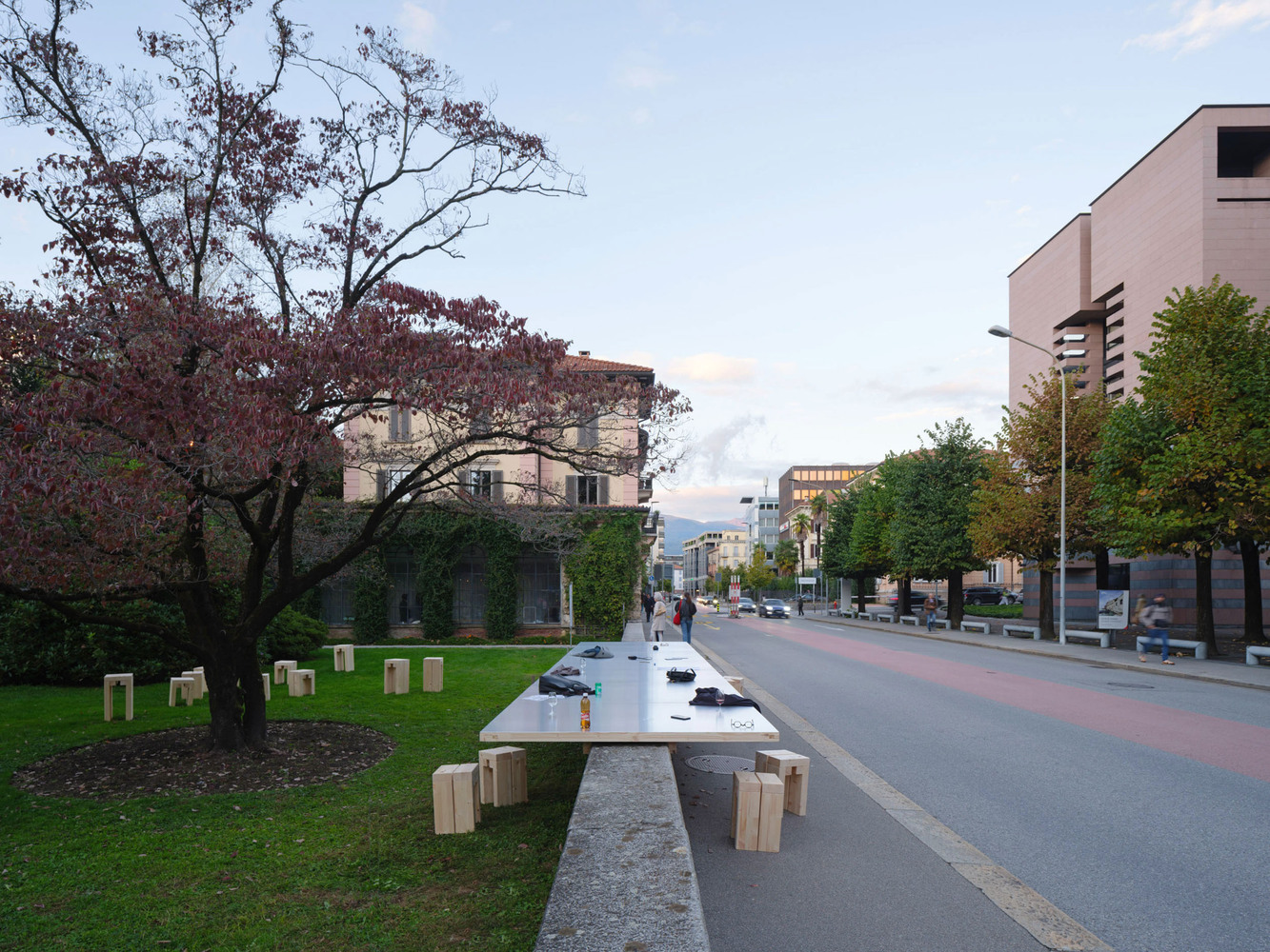 Stainless steel communal table with timber stools along a pedestrian boulevard under autumn trees at dusk