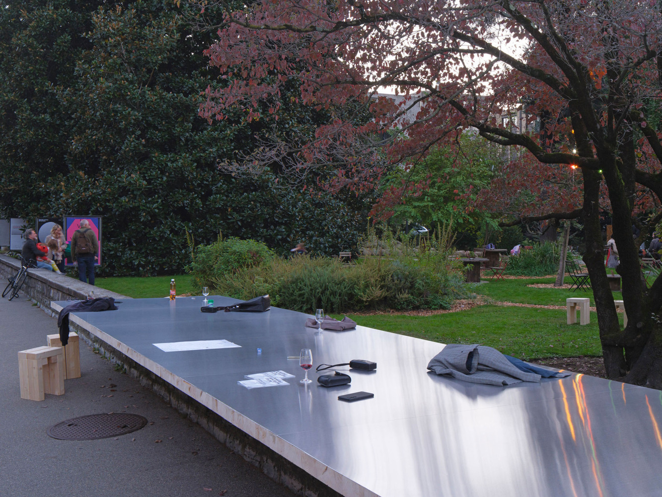 Mirrored tabletop installation with timber stools in a public park beneath branching trees at twilight