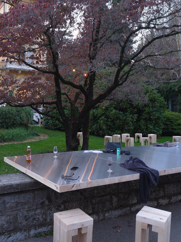 Close view of the reflective metal table surface with timber stools and wine glasses at dusk
