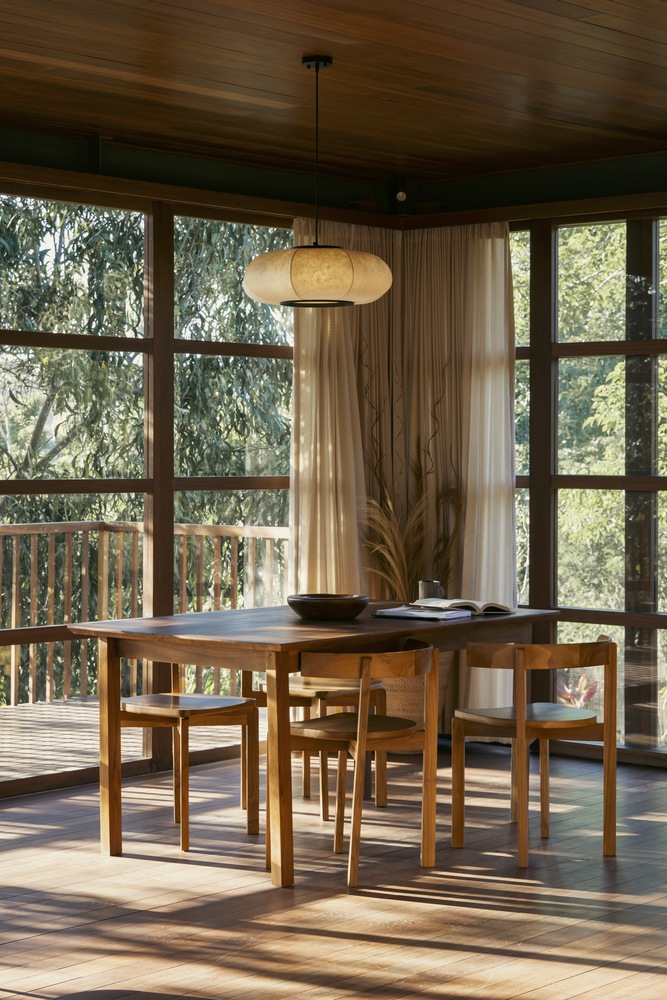 Dining table beneath a pendant light surrounded by full-height gridded windows overlooking the forest