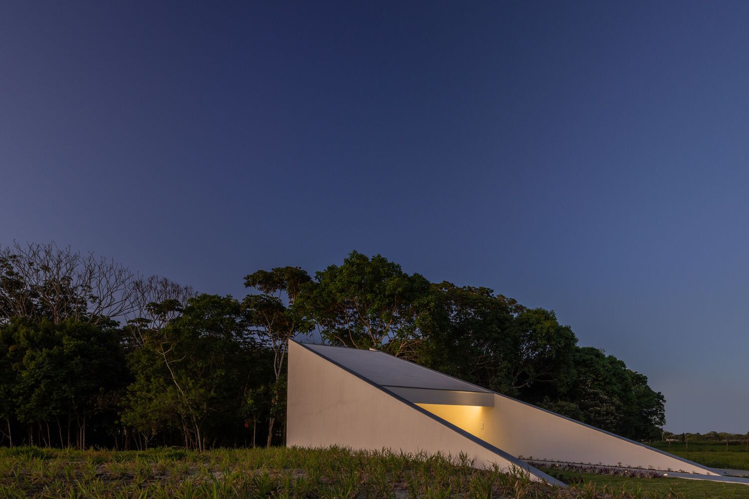 Angled roof plane with illuminated slit window photographed at dusk against the treeline