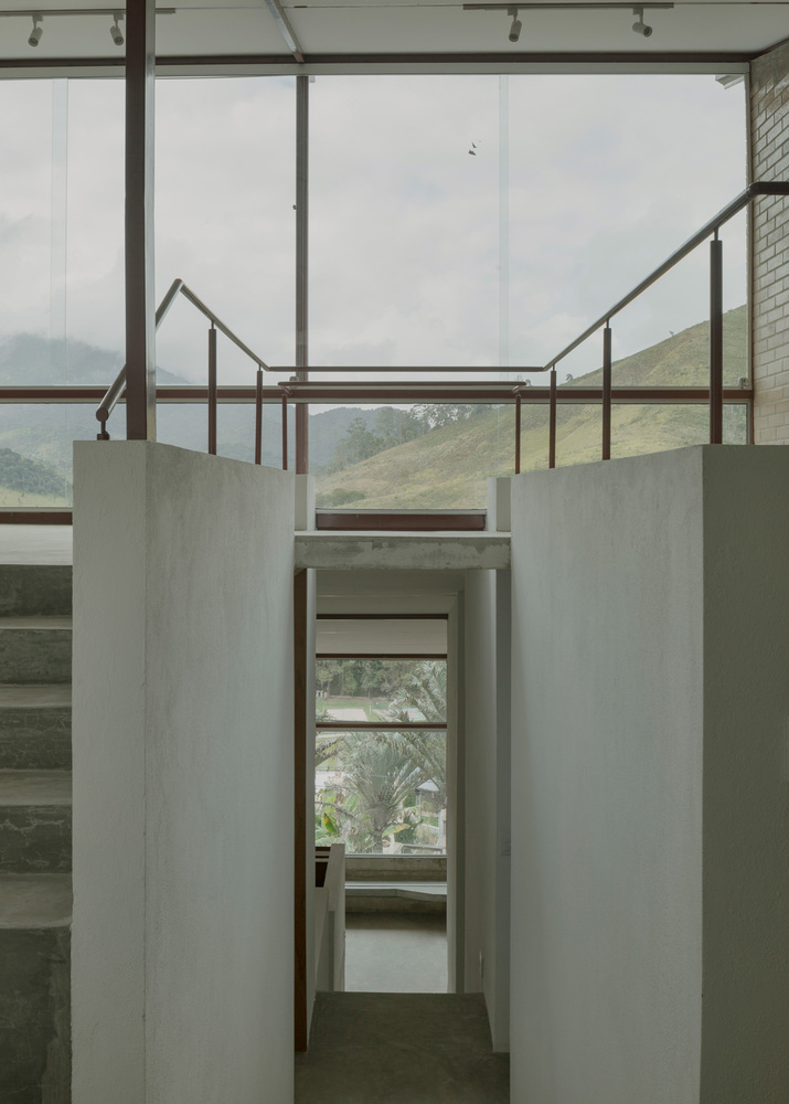 Concrete-walled corridor with steel-framed glazing above and view through to mezzanine under overcast sky