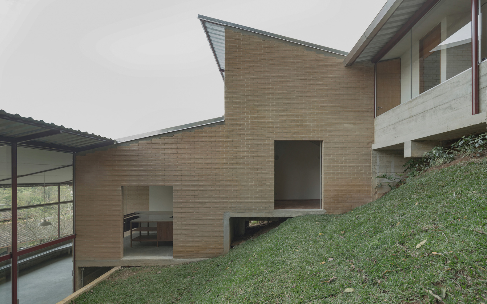 Tan brick facade with recessed windows opening onto a grass-covered hillside under overcast skies