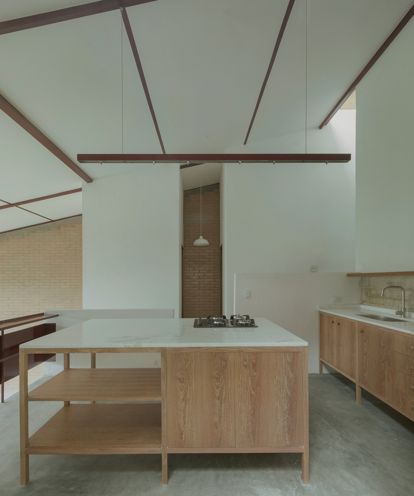 Kitchen island with timber shelving and white countertop beneath red steel beams and a double-height ceiling