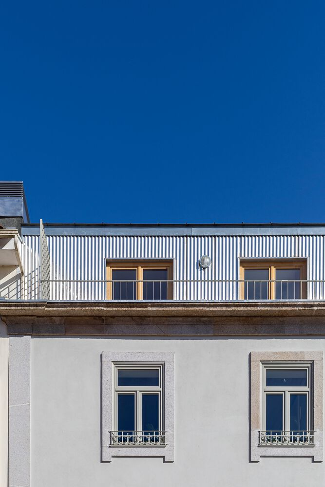 Street facade showing a corrugated metal rooftop extension above a white plastered base with framed windows under blue sky