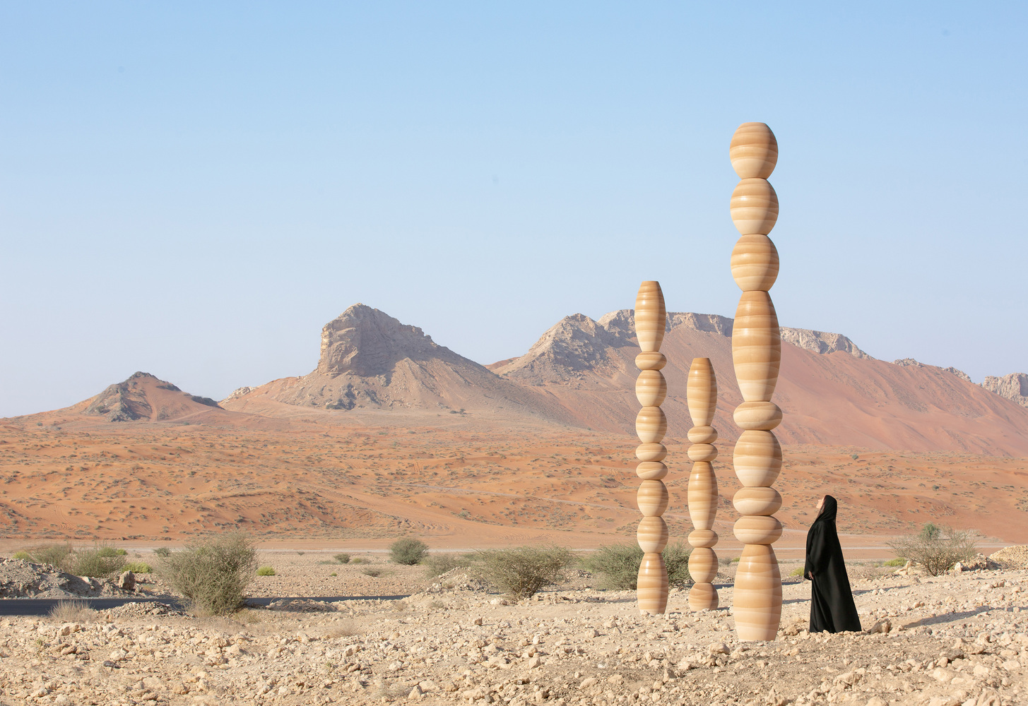 Three stacked timber columns in a desert landscape with a figure in black and distant rocky mountains