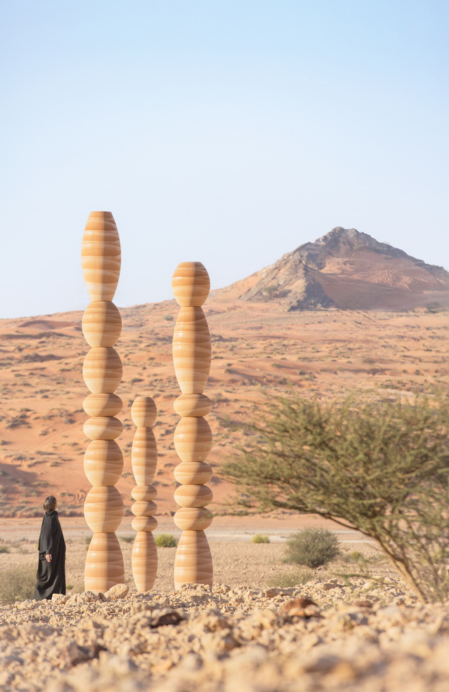 Three laminated wood columns beside a lone tree with a figure at the base and mountain beyond