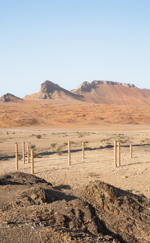 Grouped timber columns with bulbous segments anchored in the sandy ground beneath a clear sky