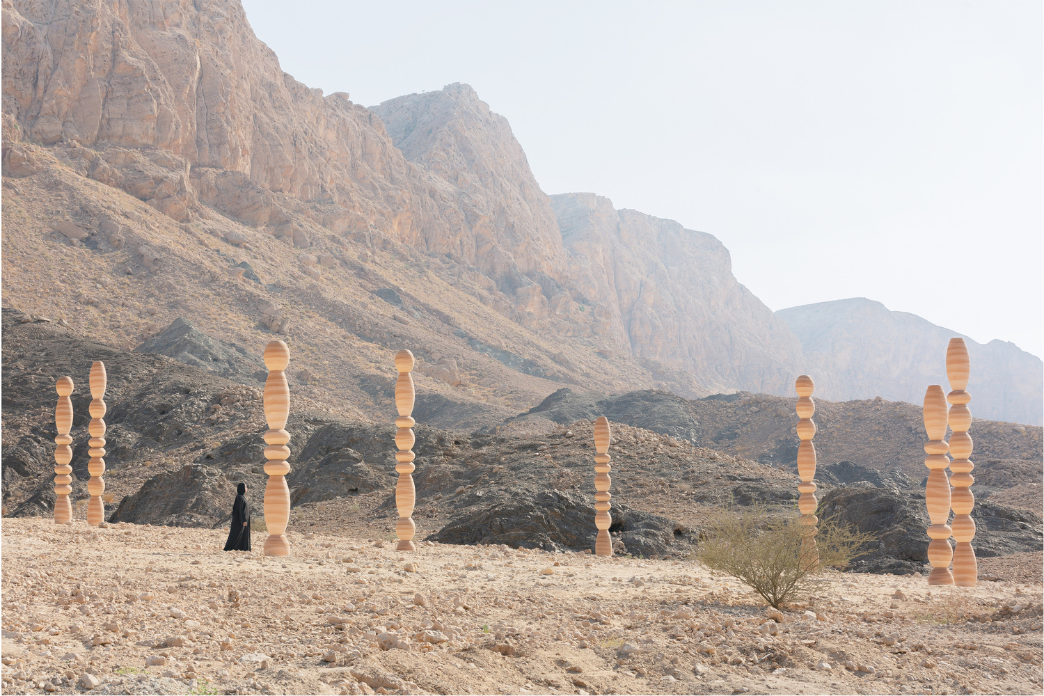 Figure in black walking past the timber columns on the stony plateau beneath towering cliffs