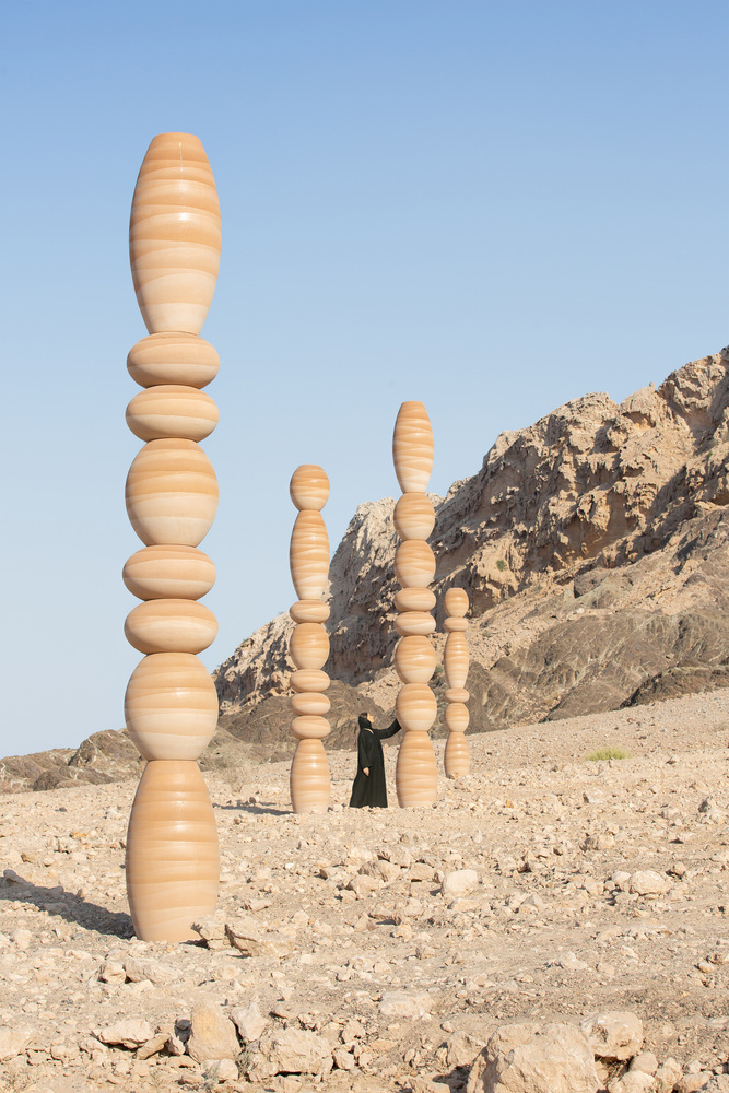 Three laminated wood columns of varying heights with a figure walking between them near rocky hills