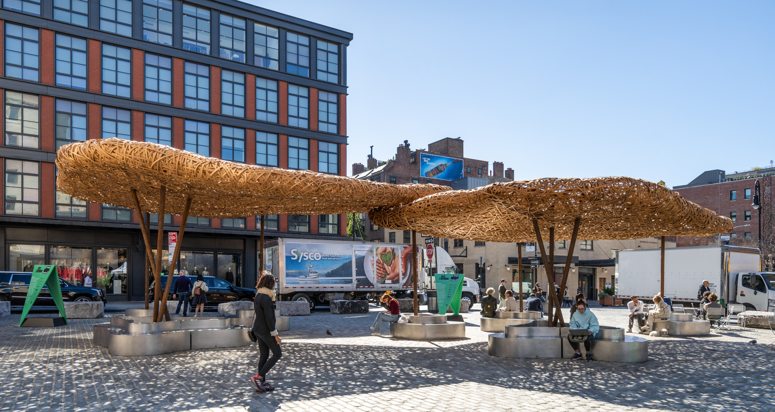 Wide view of clustered woven timber shade structures on a paved plaza with people seated at tables below