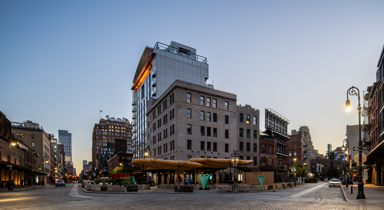 Street view of the woven timber canopies at dusk with seating and street lights along the plaza edge