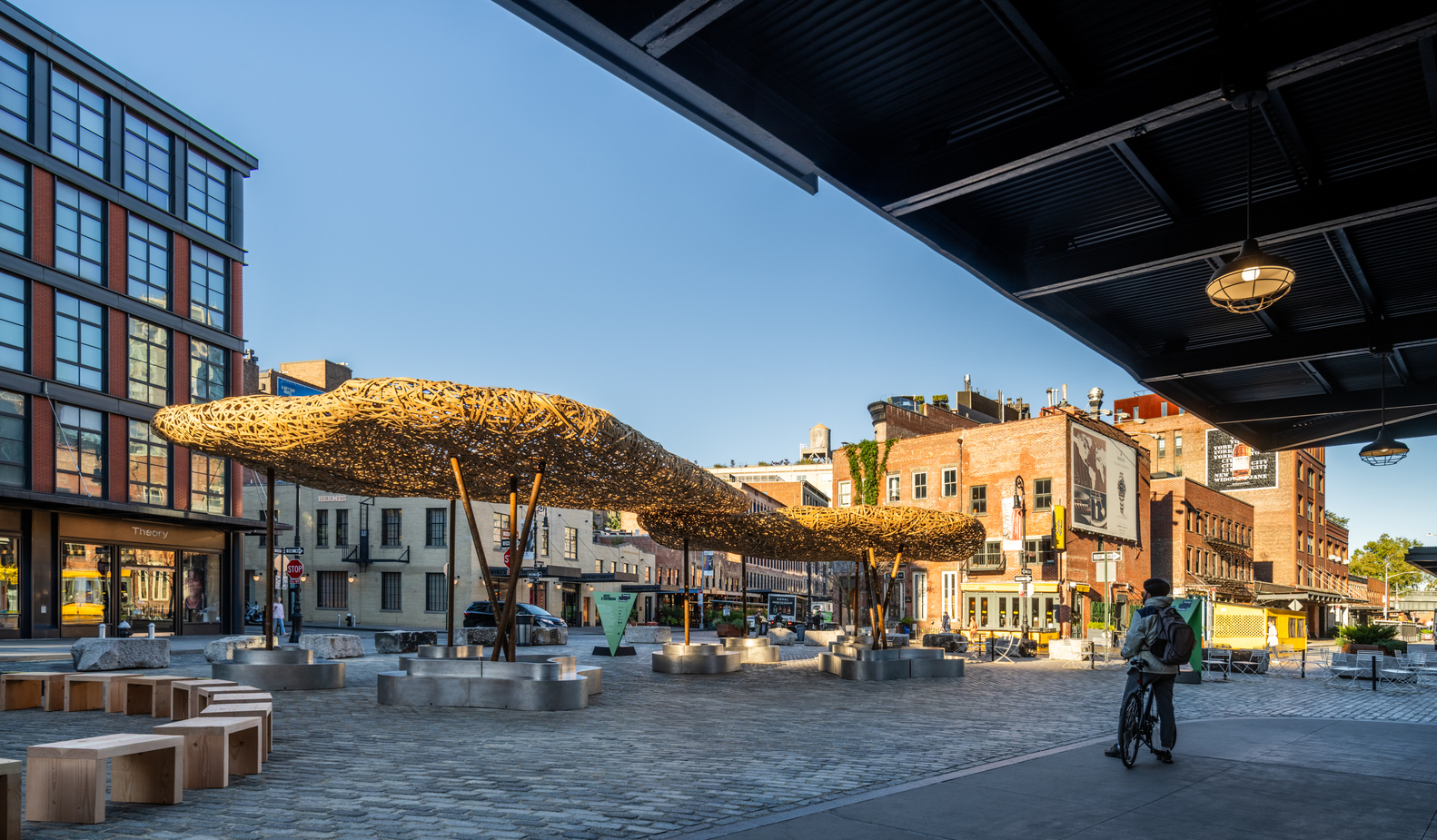 Long view of the clustered woven canopies in evening light with a cyclist crossing the plaza
