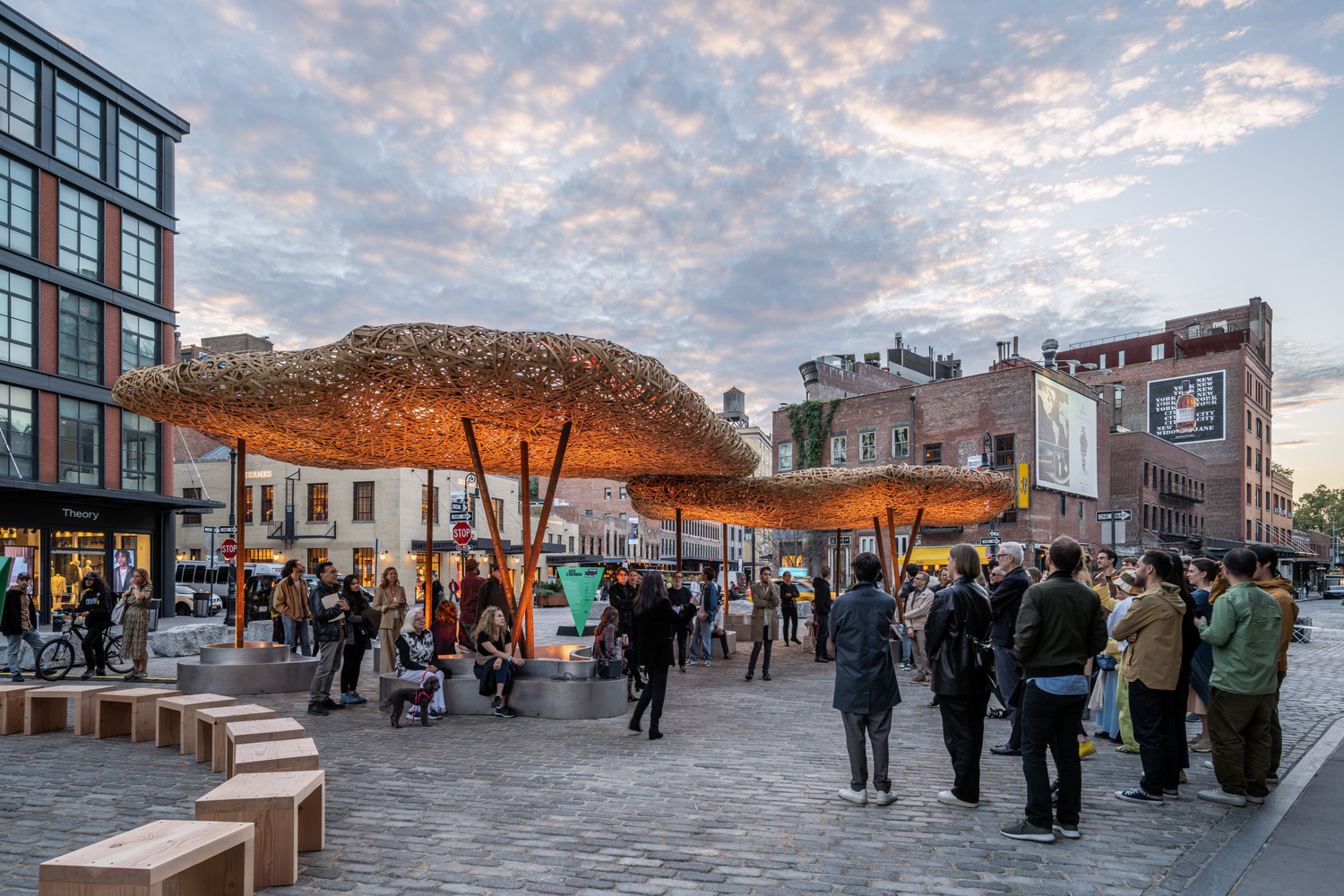 Tree-shaped woven canopy structures illuminated at dusk in a public plaza with gathered visitors
