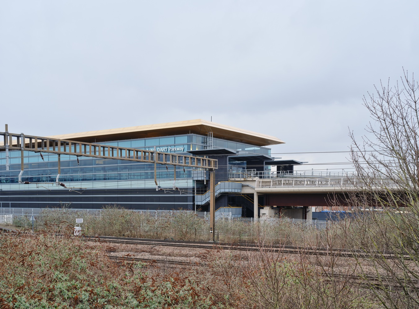Glass-clad terminal with elevated walkway bridge viewed from trackside with dormant plants