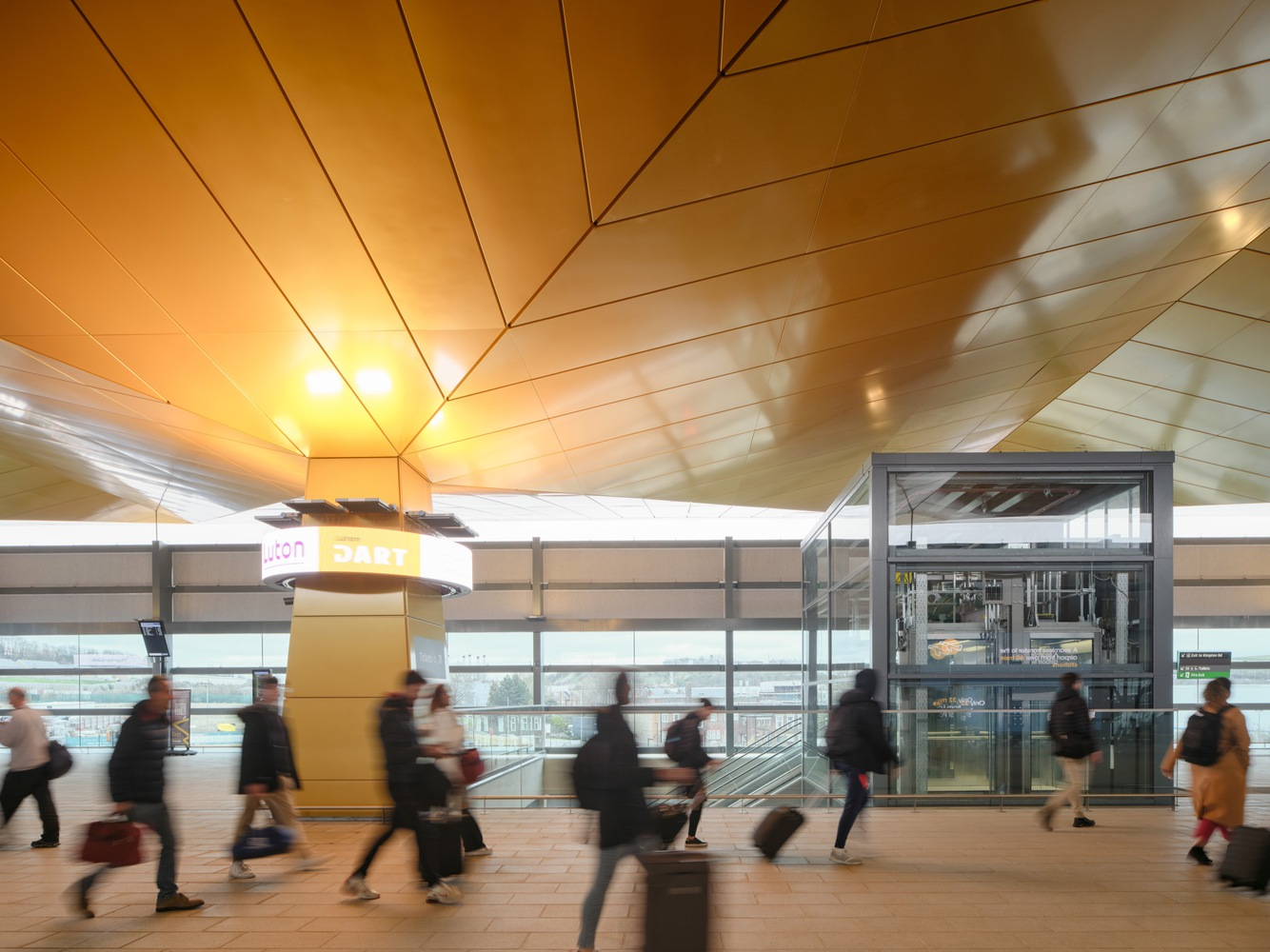 Transit hall with golden paneled ceiling and travelers with luggage passing near glass elevator shaft