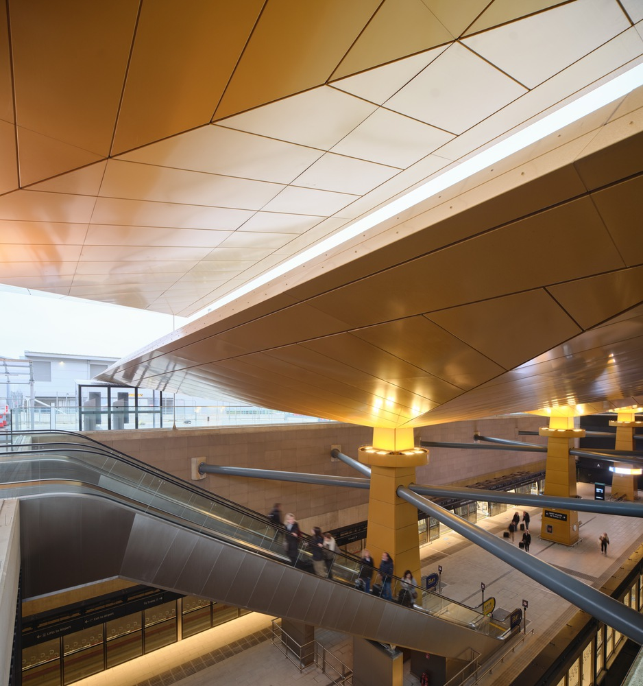 Multilevel atrium with escalators, golden ceiling planes, and commuters moving between platform levels