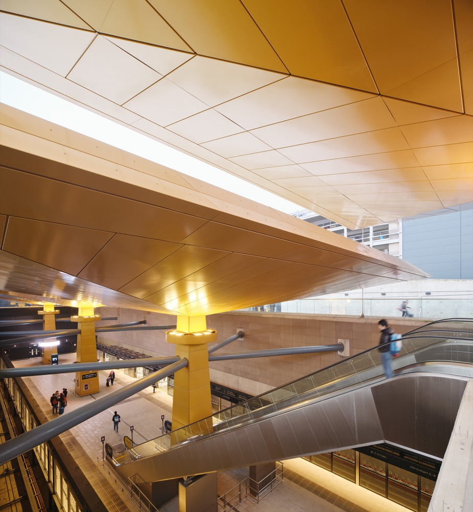 View across atrium showing layered golden ceiling, yellow columns, and blurred figures on escalators