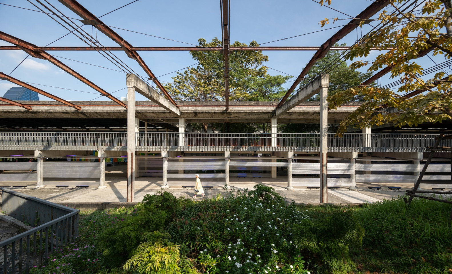 Covered outdoor platform with concrete columns and planted beds beneath a metal truss canopy