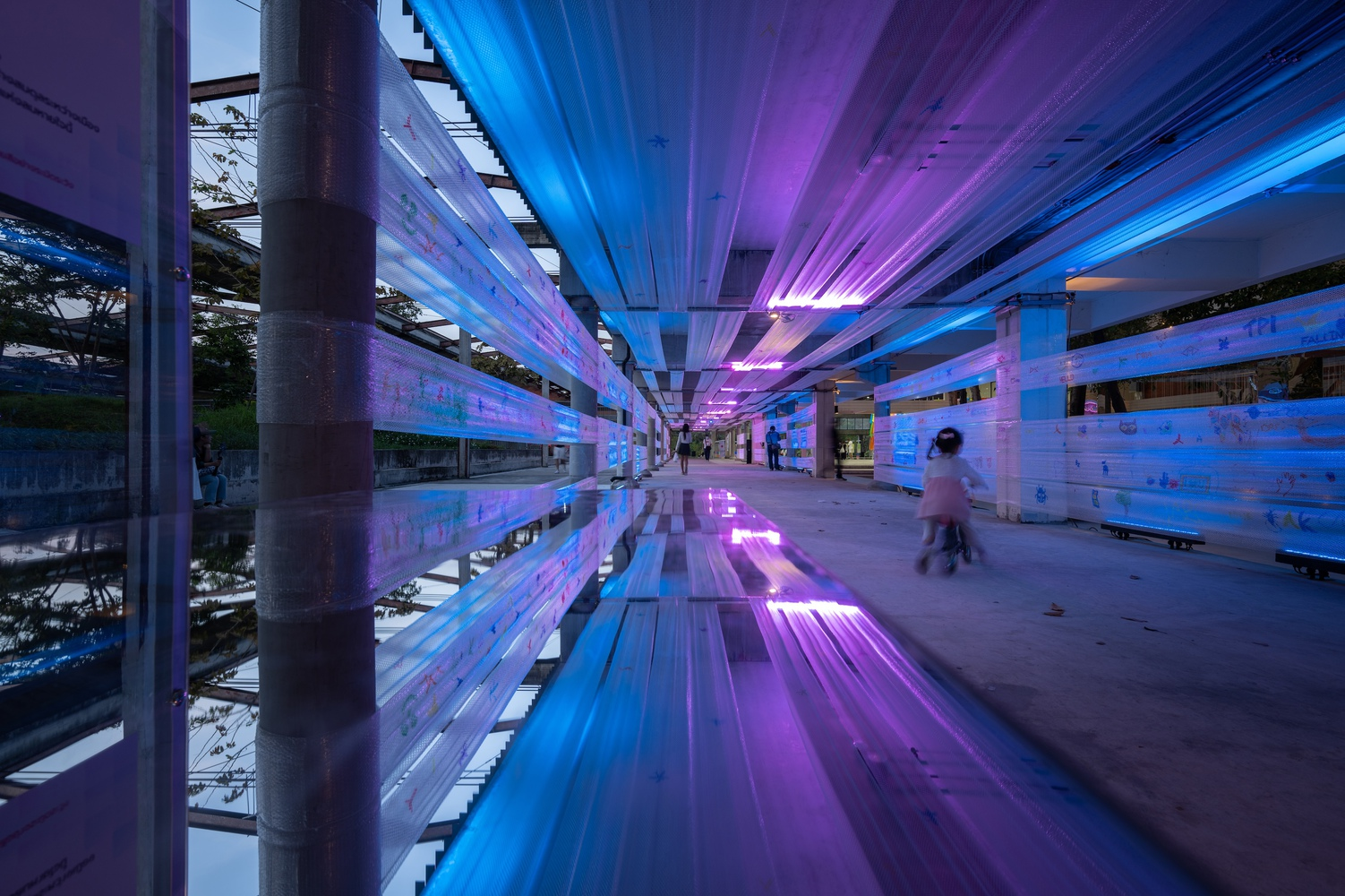 Covered corridor with reflective floor illuminated by purple and blue light strips at dusk