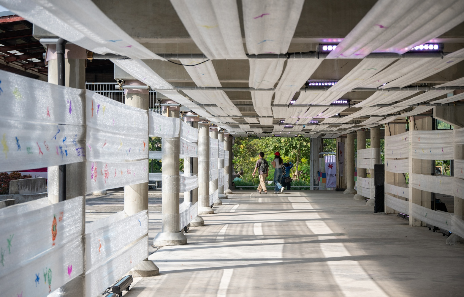 Covered walkway with radiating pleated ceiling panels, white fabric screens, and cylindrical columns as pedestrians pass through
