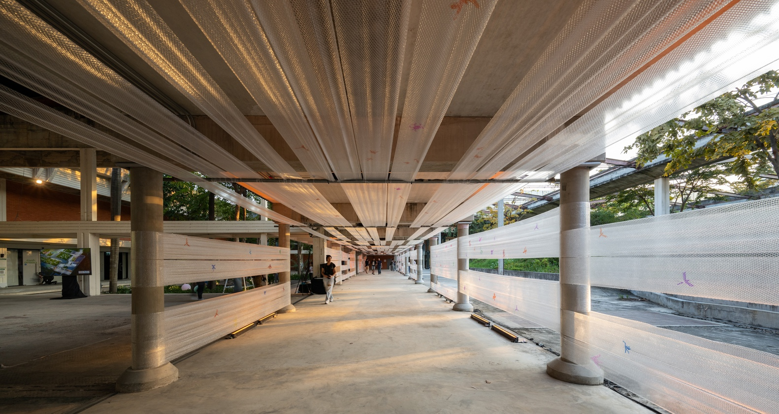 Corridor at dusk with warm interior lighting visible through glazing and the pleated ceiling overhead