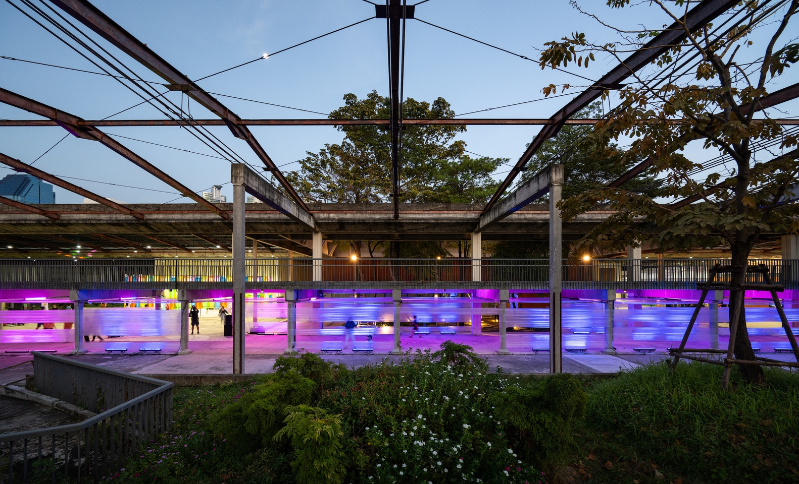 Central courtyard with steel truss canopy above magenta-illuminated screening and planted landscape at dusk