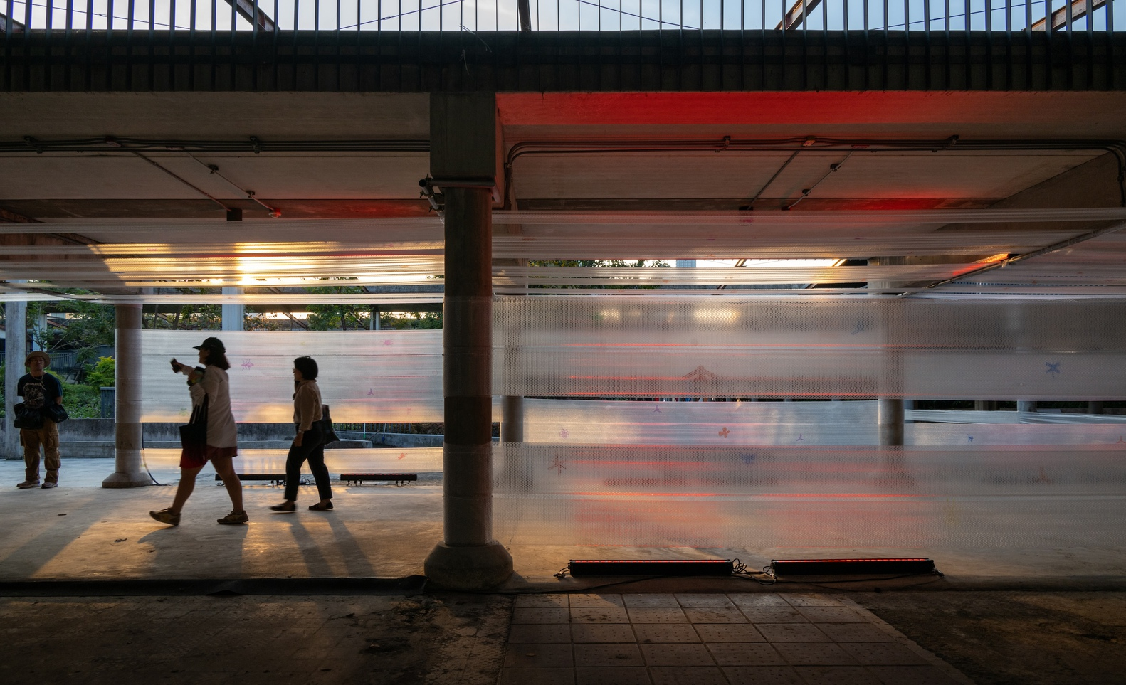 Platform edge with concrete columns and pedestrians walking past translucent backlit wall panels at dusk