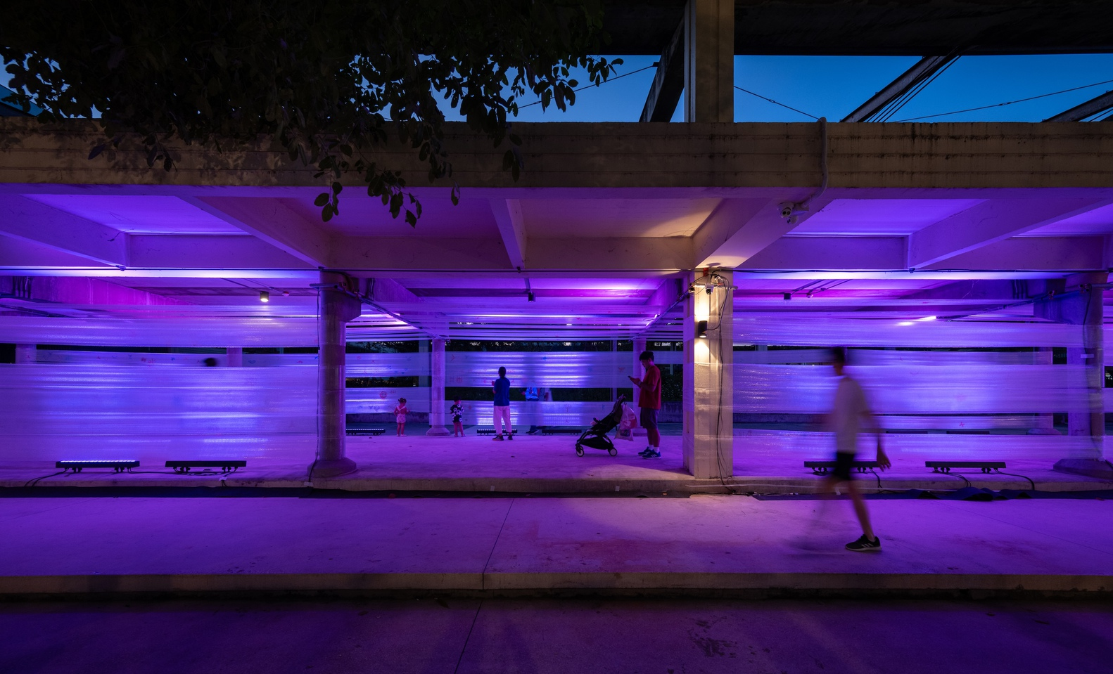 Covered pedestrian walkway with purple LED lighting along concrete columns and a person in motion at dusk