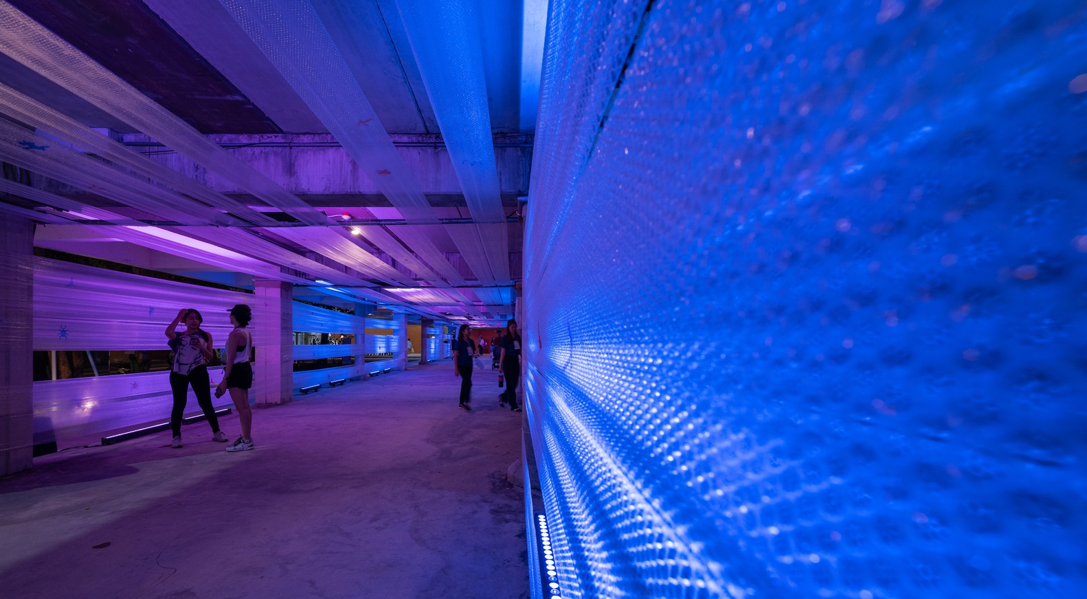 Close-up of blue LED screen wall with pixelated pattern and visitors beneath purple-lit concrete ceiling