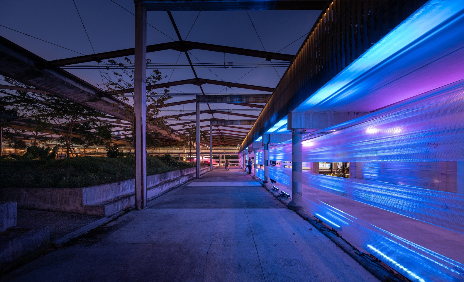 Linear walkway with steel truss canopy and illuminated blue facade wall at twilight