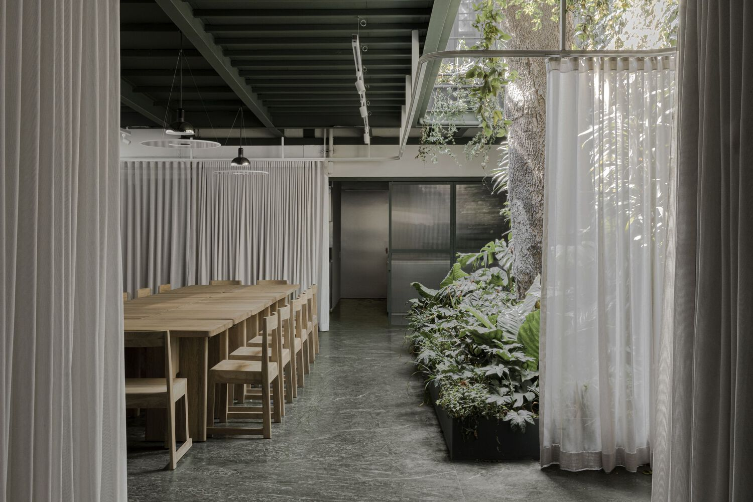 Dining area with timber table and stools framed by sheer curtains and an indoor planted bed