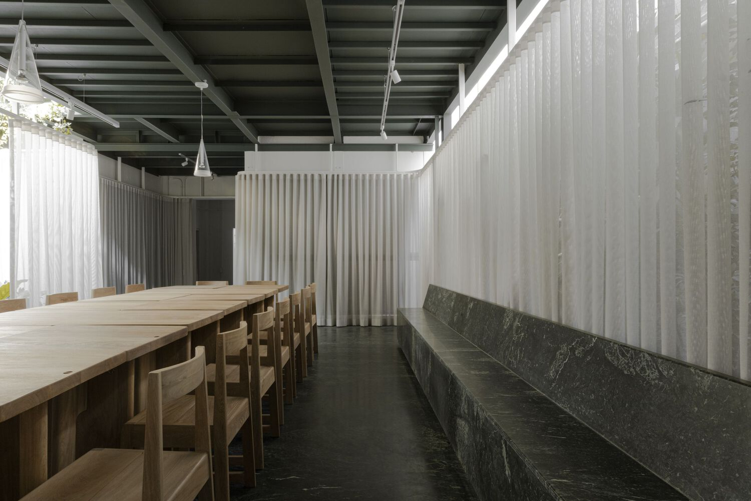 Long timber communal table beside a dark stone bench with sheer curtain partitions and clerestory windows
