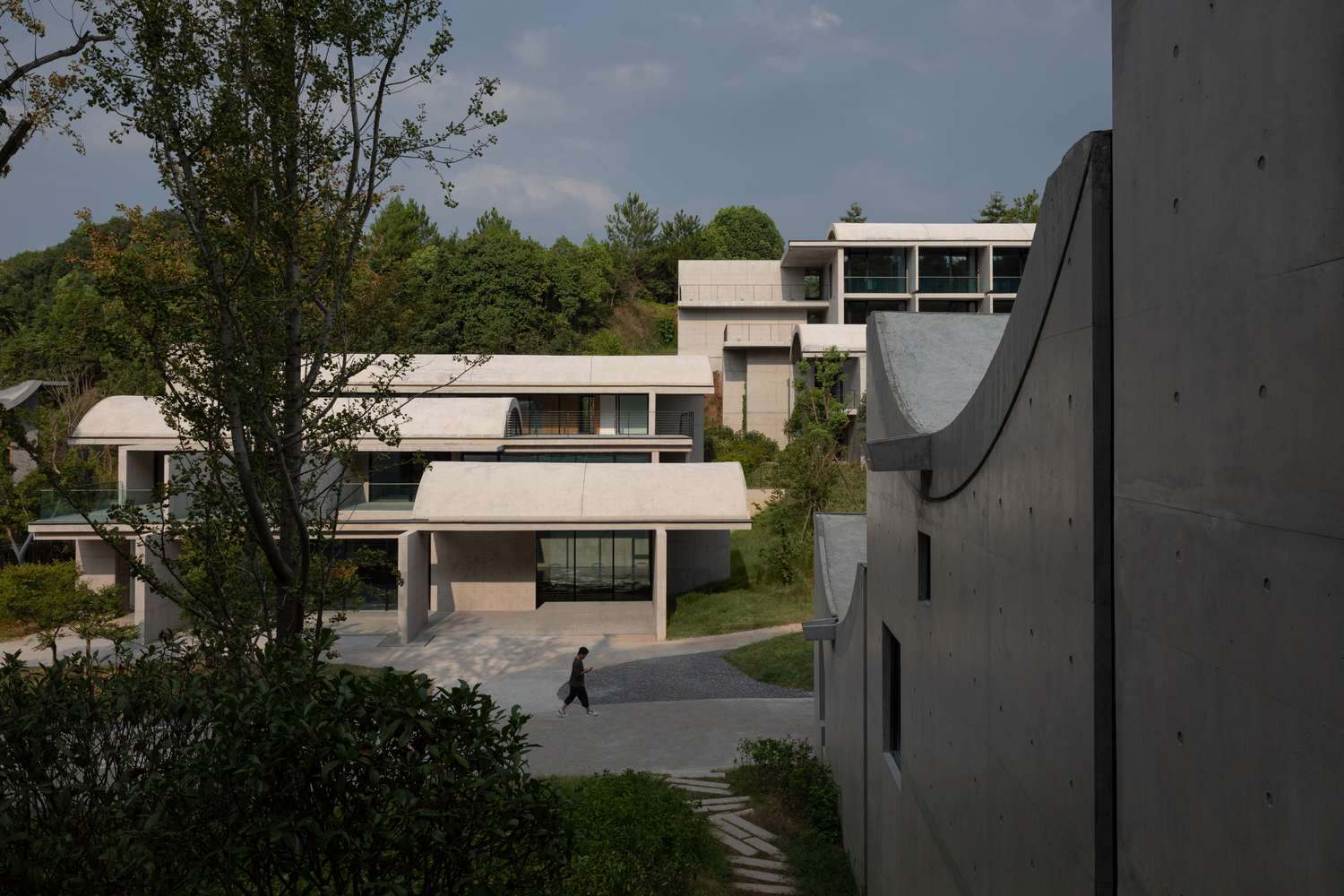 Complex of white concrete volumes with curved roofs terracing up a wooded hillside with a figure crossing the plaza