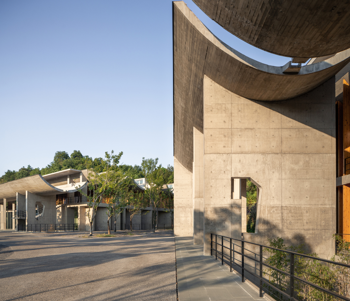 Curved concrete canopy with board-formed soffit extending over a paved courtyard in afternoon sunlight