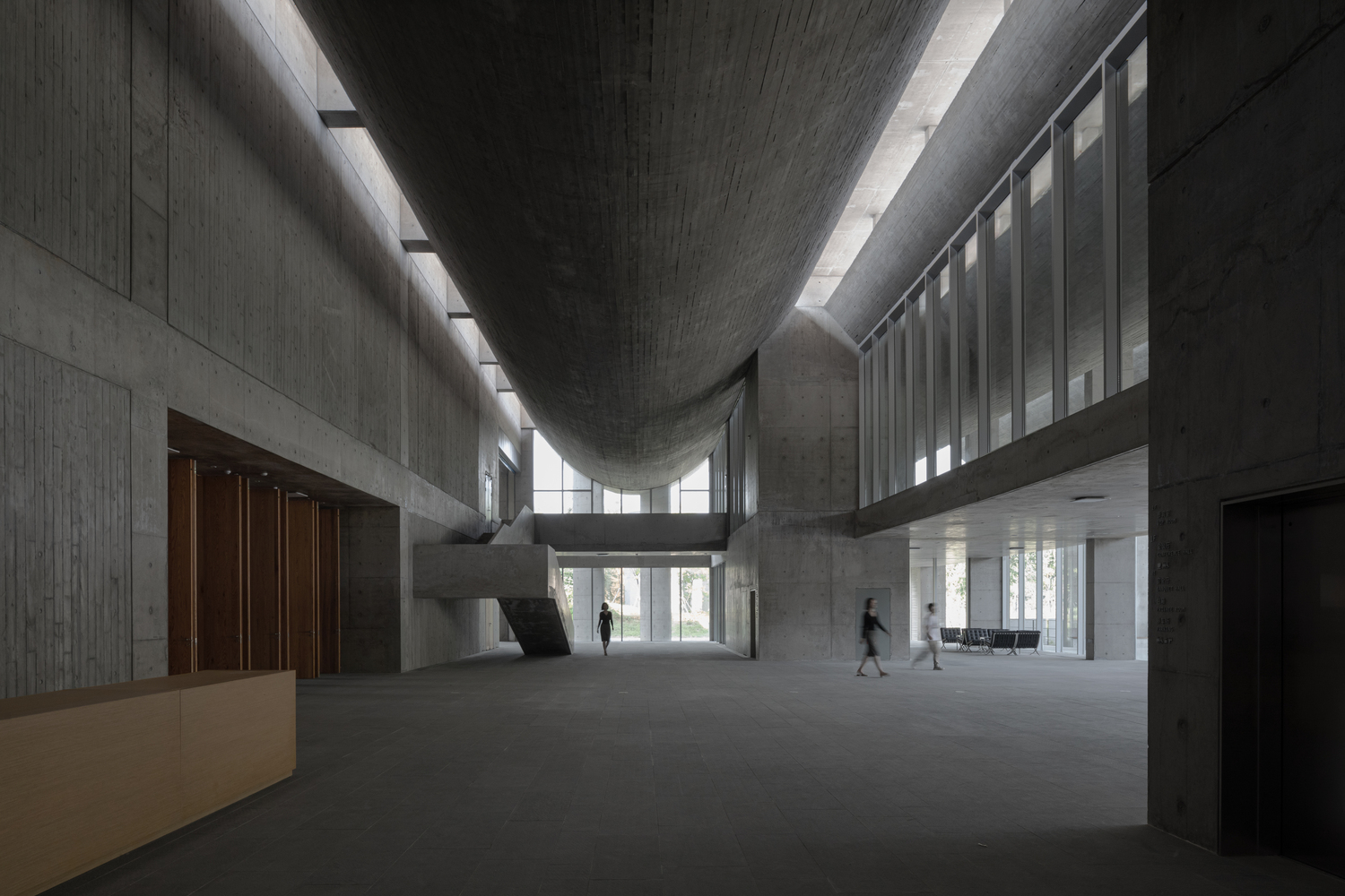 Double-height lobby with curved concrete ceiling vault and clerestory windows illuminating figures crossing the floor