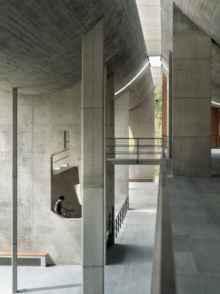 Multi-level interior with board-formed concrete columns and spiral staircase bathed in natural light from skylights above