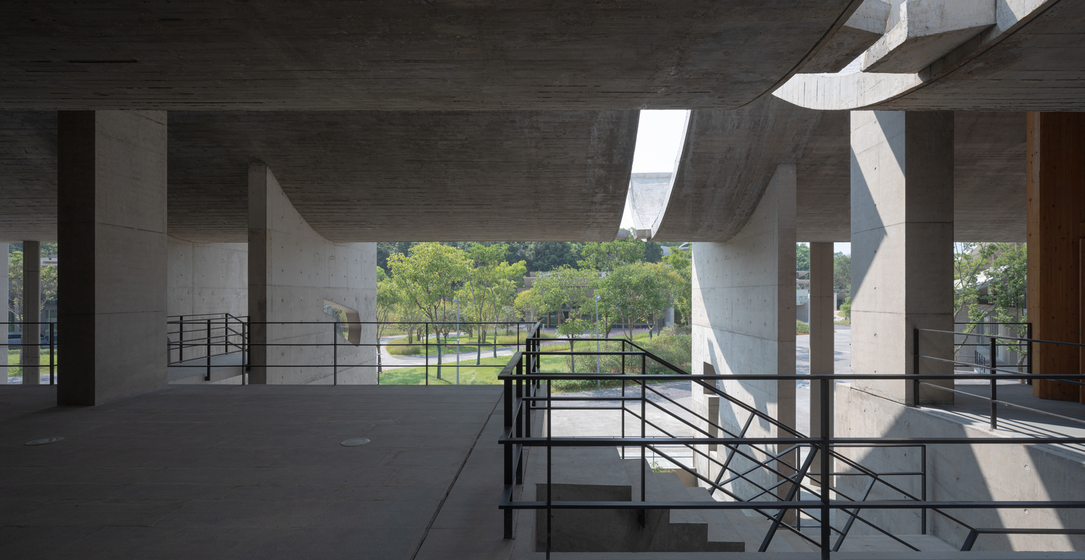 Open concrete space with metal railings and angular ceiling apertures framing a view of the landscaped grounds