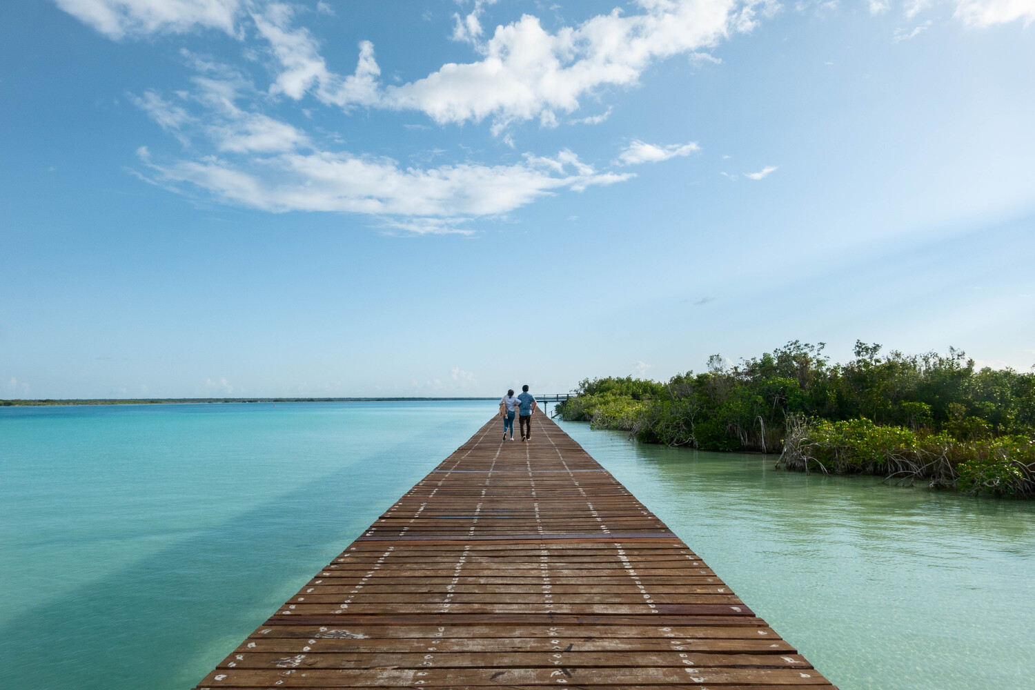 Weathered timber boardwalk extending across turquoise lagoon waters toward mangrove vegetation with two visitors walking
