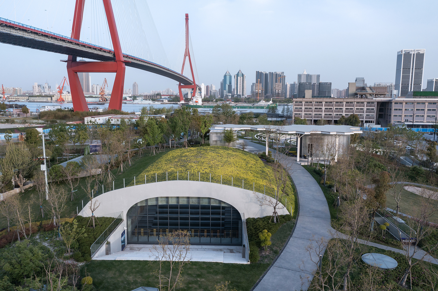 Grass-covered roof arching over glazed facade with cable-stayed bridge and river skyline in background